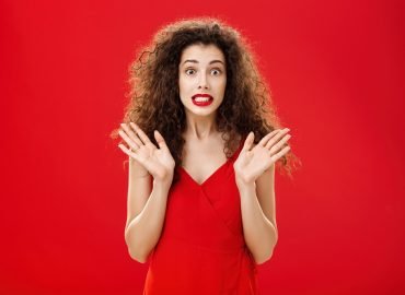 Woman feeling nervous and concerned messing up important papers clenching teeth and popping eyes being worried and troubled waving raised palms in clueless and perplexed gesture over red background. Copy space